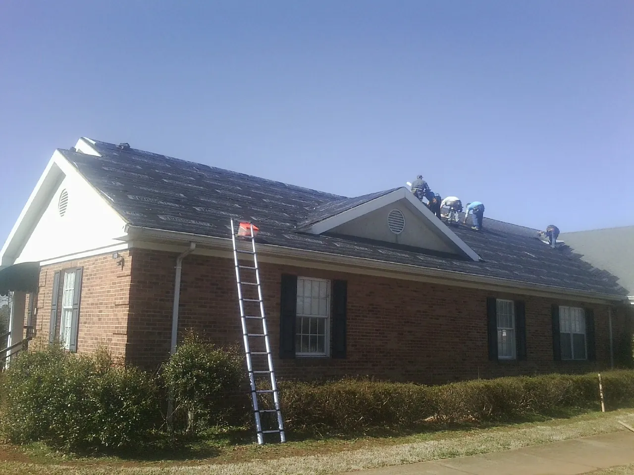 Crew tearing shingles off a brick ranch roof with a ladder set — Emergency Roof Repair work by our crew in Sandy Ridge