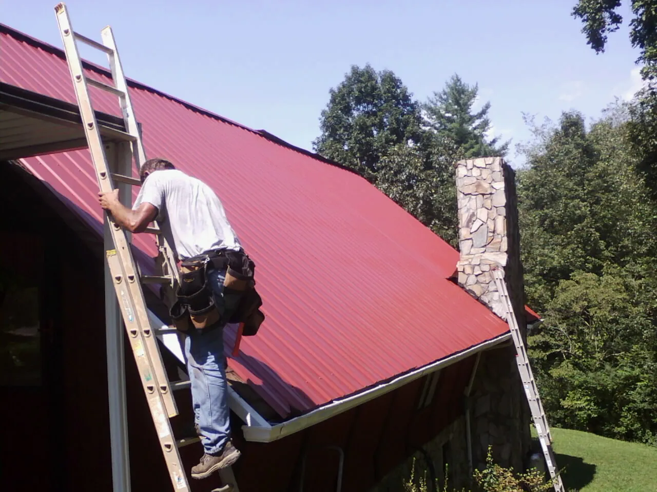 Roofer on a ladder beside a red metal roof and stone chimney — Soffit & Fascia Repair by Roofing Brothers in Sandy Ridge
